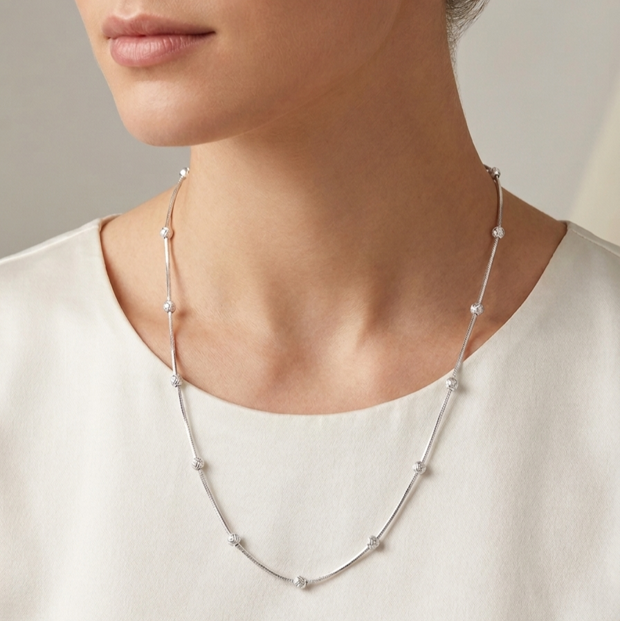 A close-up of a model wearing a delicate silver necklace with small, evenly spaced round clear stones, styled with a simple cream-colored top.