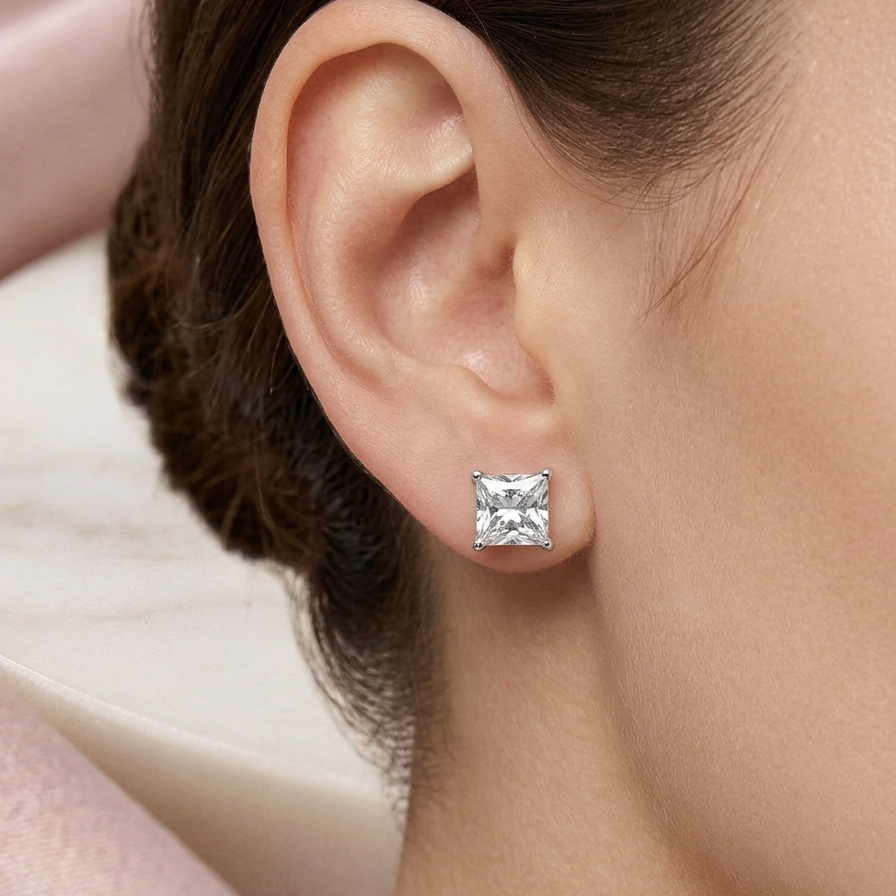 A close-up of a model's ear wearing a single princess-cut clear gemstone stud earring in a silver setting.