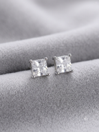 A pair of princess-cut clear gemstone stud earrings in a silver setting, displayed on a textured gray velvet surface.