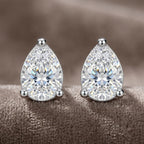 A pair of pear-cut diamond stud earrings in a silver-toned setting, displayed on a soft, textured brown surface.
