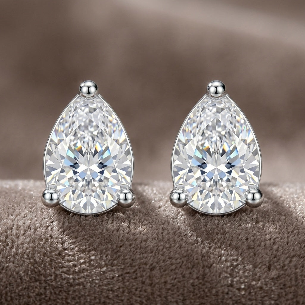 A pair of pear-cut diamond stud earrings in a silver-toned setting, displayed on a soft, textured brown surface.