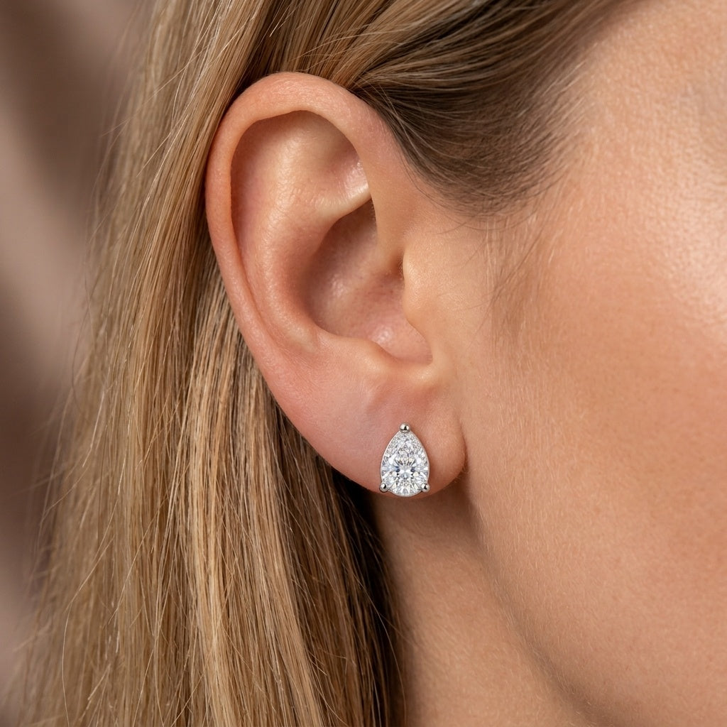 A close-up of a woman's ear wearing a small pear-shaped stud earring with a clear gemstone in a silver-toned setting.
