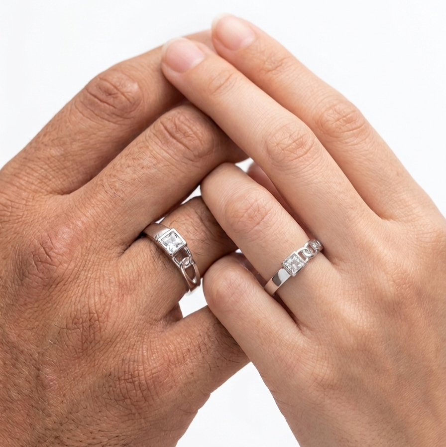 Two hands clasped together, each wearing a silver band ring with a small clear gemstone, photographed against a plain white background.