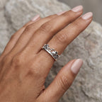 A silver ring with a detailed animal silhouette pattern, worn on a hand with light pink manicured nails, set against a blurred gray stone background.
