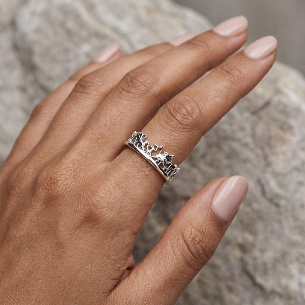 A silver ring with a detailed animal silhouette pattern, worn on a hand with light pink manicured nails, set against a blurred gray stone background.