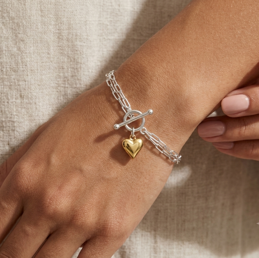 A close-up of a person's wrist wearing a delicate silver chain bracelet with a toggle clasp and a small dangling gold heart charm, set against a neutral beige linen background.