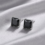 A pair of square stud earrings with faceted black gemstones in silver-toned prong settings, displayed on a draped silver satin fabric.