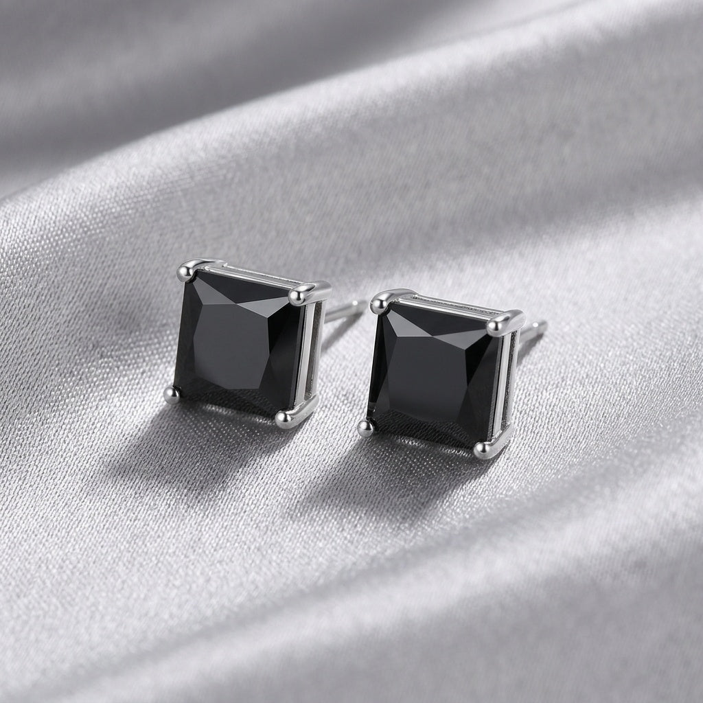 A pair of square stud earrings with faceted black gemstones in silver-toned prong settings, displayed on a draped silver satin fabric.