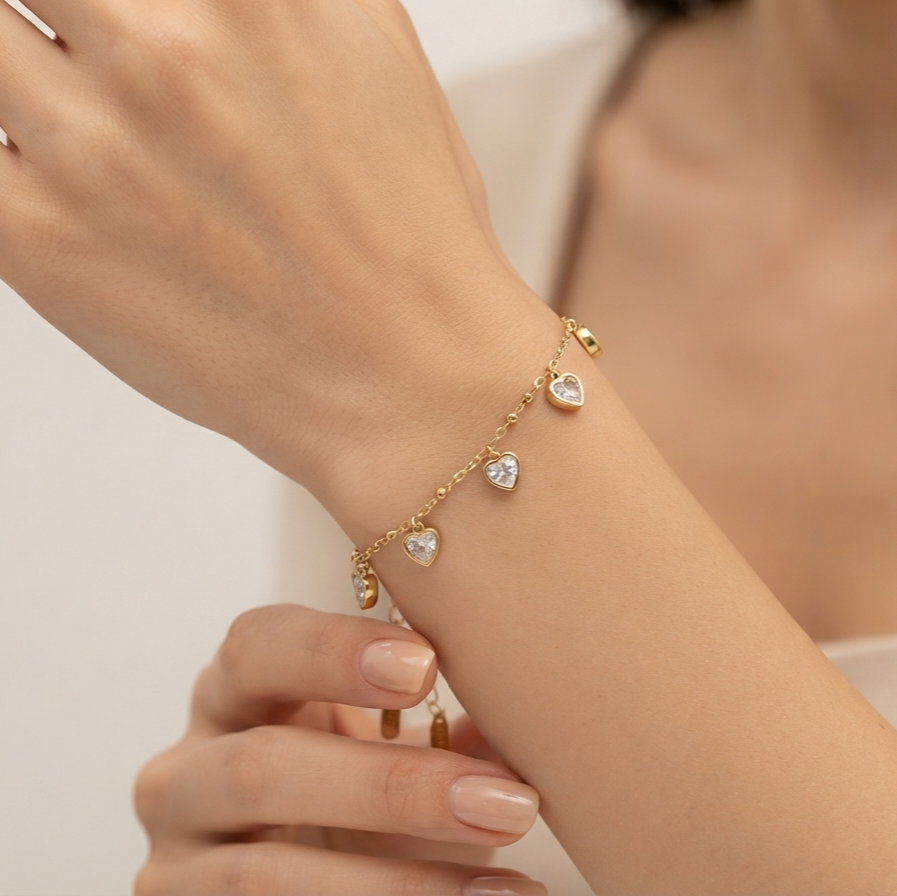 A delicate gold chain bracelet with three heart-shaped crystal charms worn on a woman's wrist, shown in a close-up lifestyle shot.