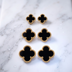 Six black clover-shaped jewelry pieces with textured gold borders, arranged on a white marble surface.