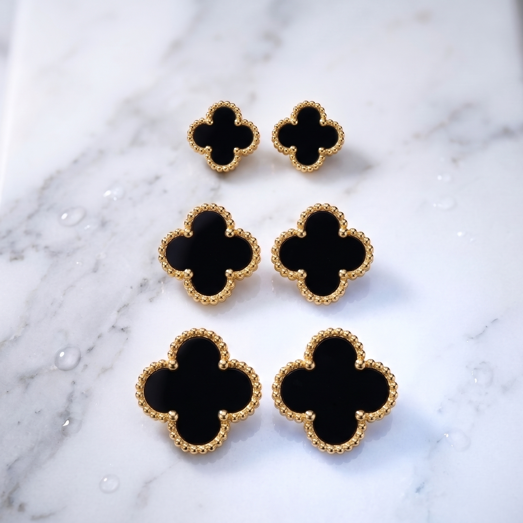 Six black clover-shaped jewelry pieces with textured gold borders, arranged on a white marble surface.
