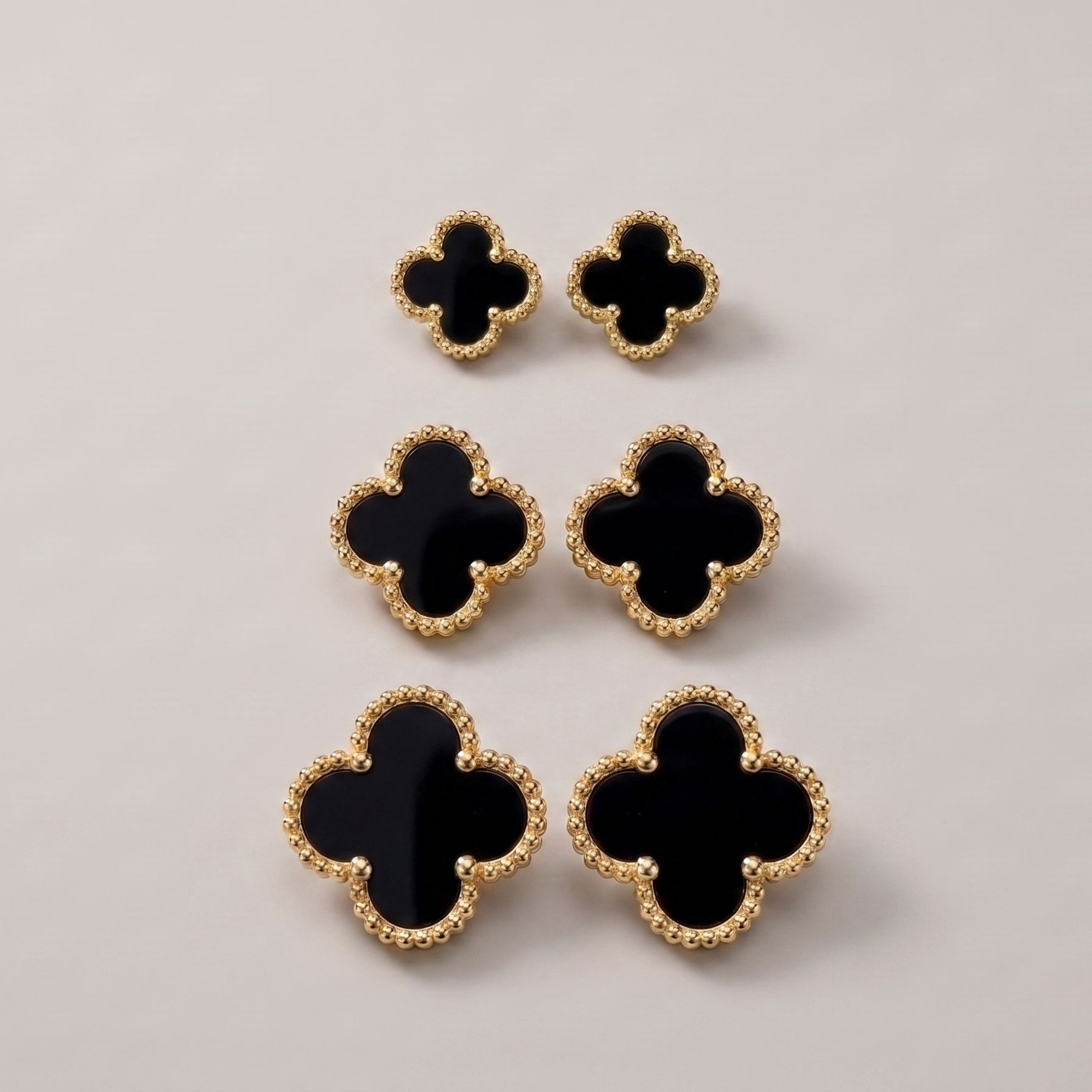 Six black clover-shaped earrings with textured gold borders, arranged in a vertical stack on a light gray background.