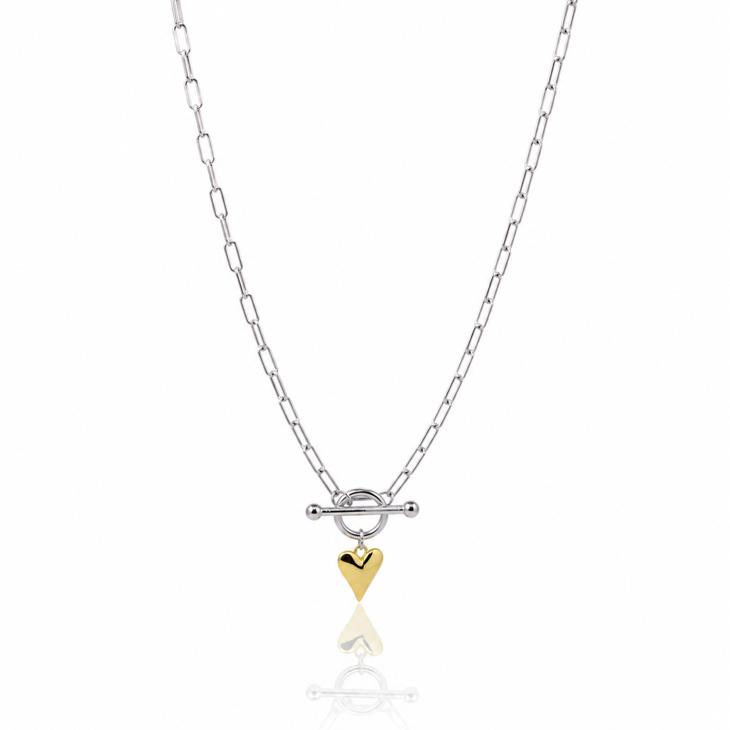 A silver paperclip chain necklace with a toggle clasp and a small dangling gold heart charm, shown on a clean white background with a soft reflection below.