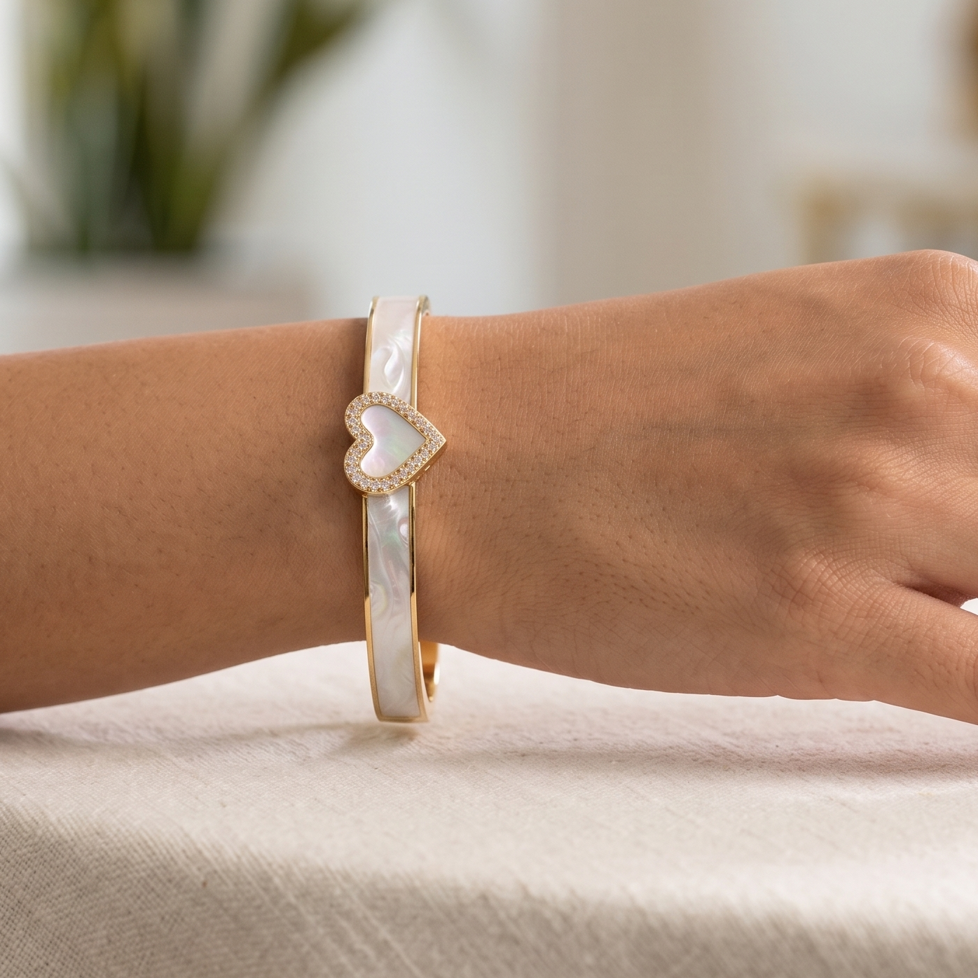 A close-up of a person's hand resting on a light beige tablecloth, wearing a gold and white beaded bracelet with a heart-shaped charm.