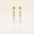 A pair of gold dangle earrings with a spherical stud, an open diamond-shaped link, and a hanging pearl, displayed on a clean white reflective surface.