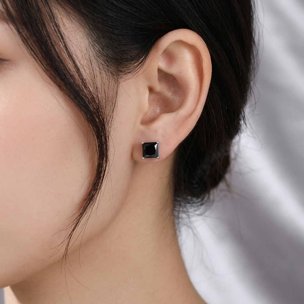 A close-up of a person's ear wearing a small square stud earring with a black center and silver-toned border.