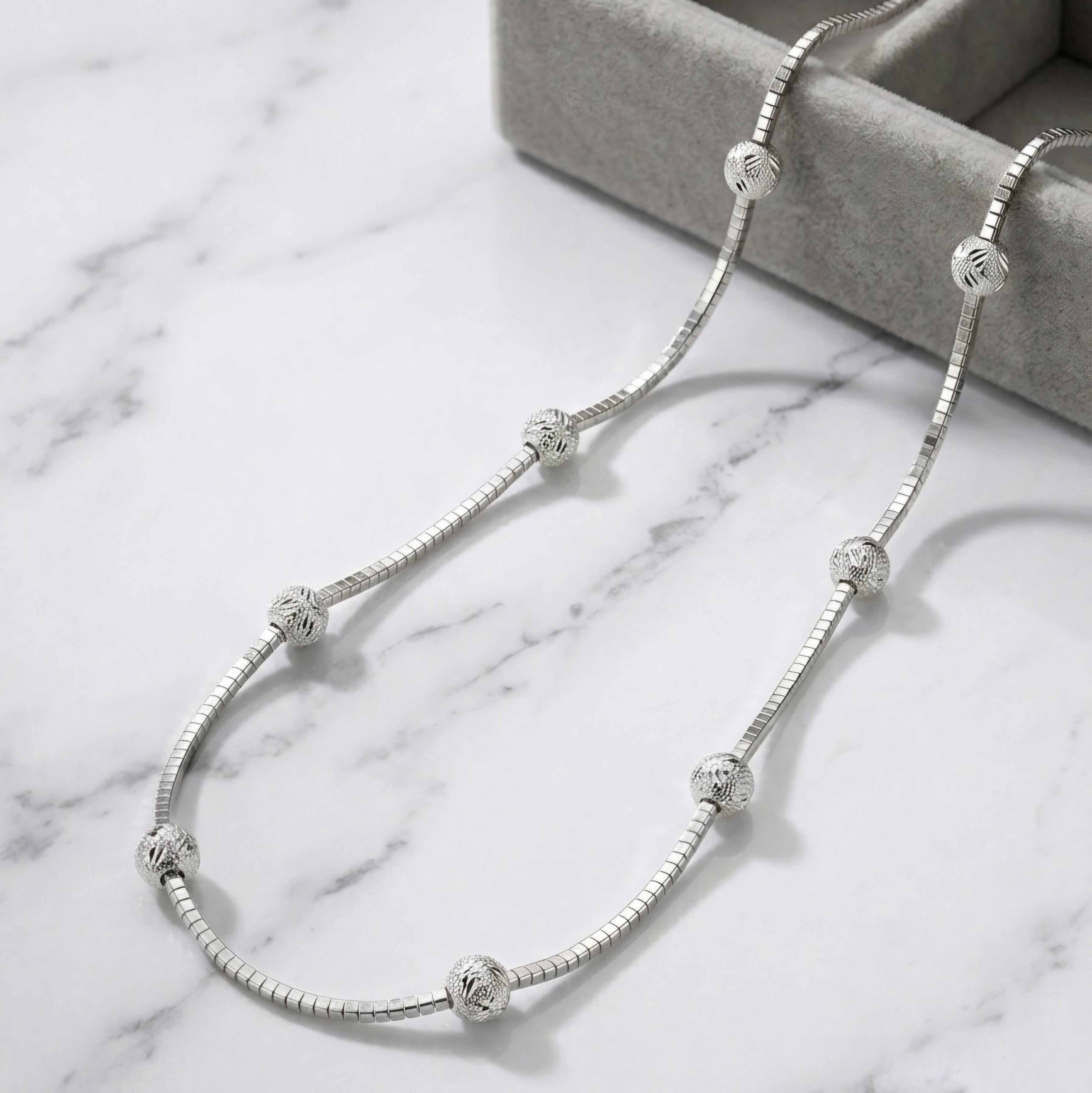 A silver snake chain necklace with textured spherical beads, displayed on a white marble surface next to a gray jewelry box.
