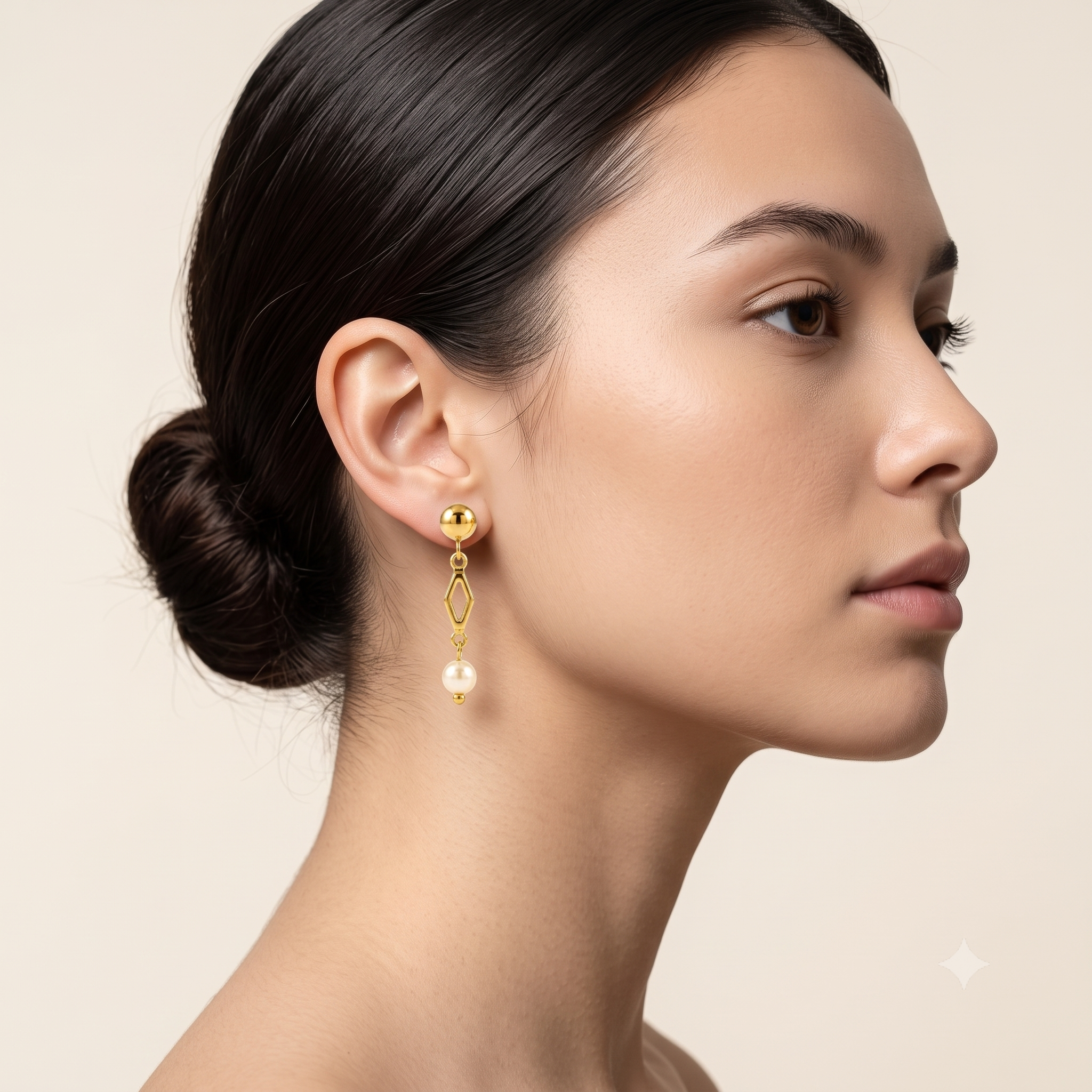 A close-up side profile of a woman with dark hair in a bun, wearing a gold dangle earring with a small white pearl drop against a soft beige background.