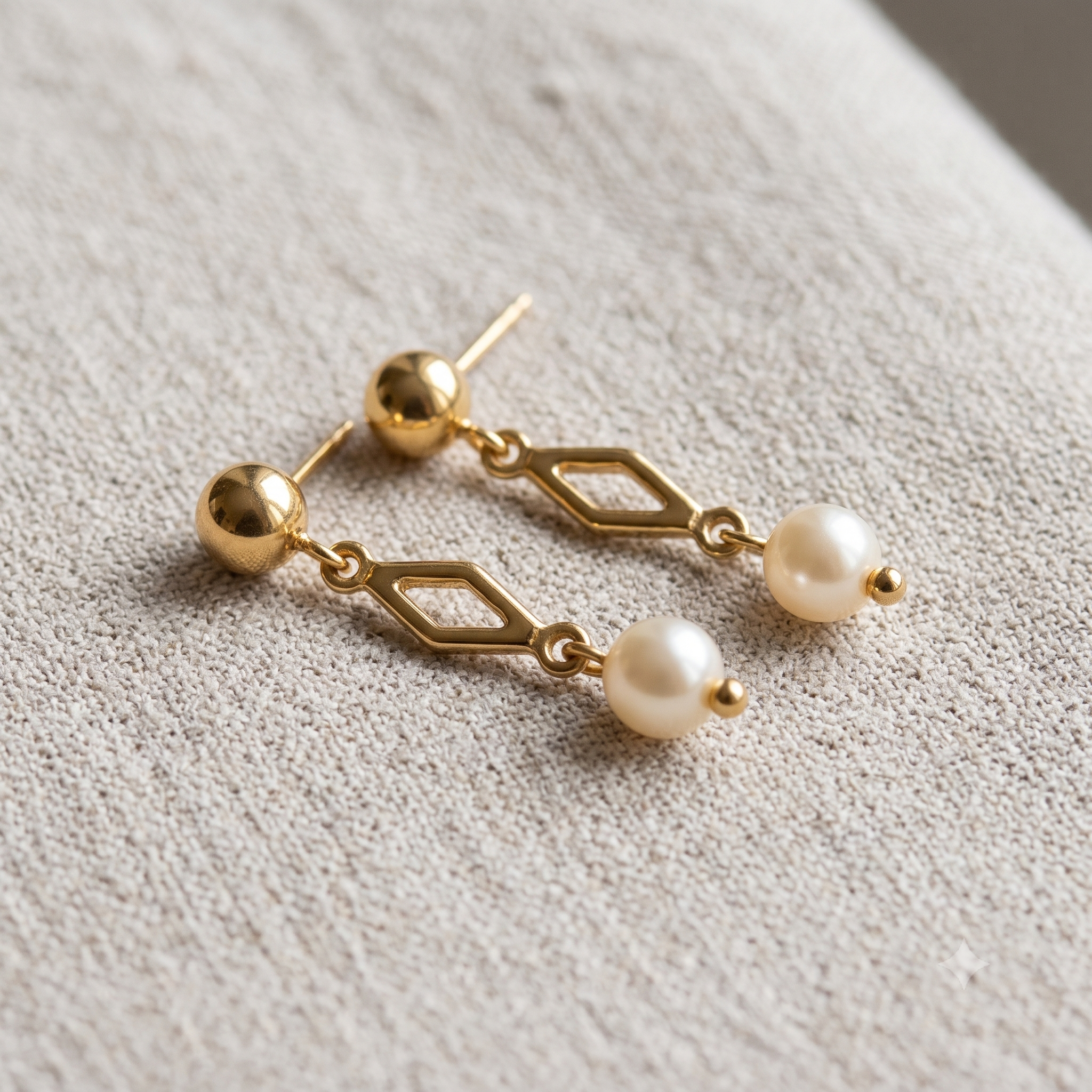 A pair of gold drop earrings with a geometric diamond-shaped link, a small gold bead, and a white pearl dangle, resting on a textured beige fabric background.