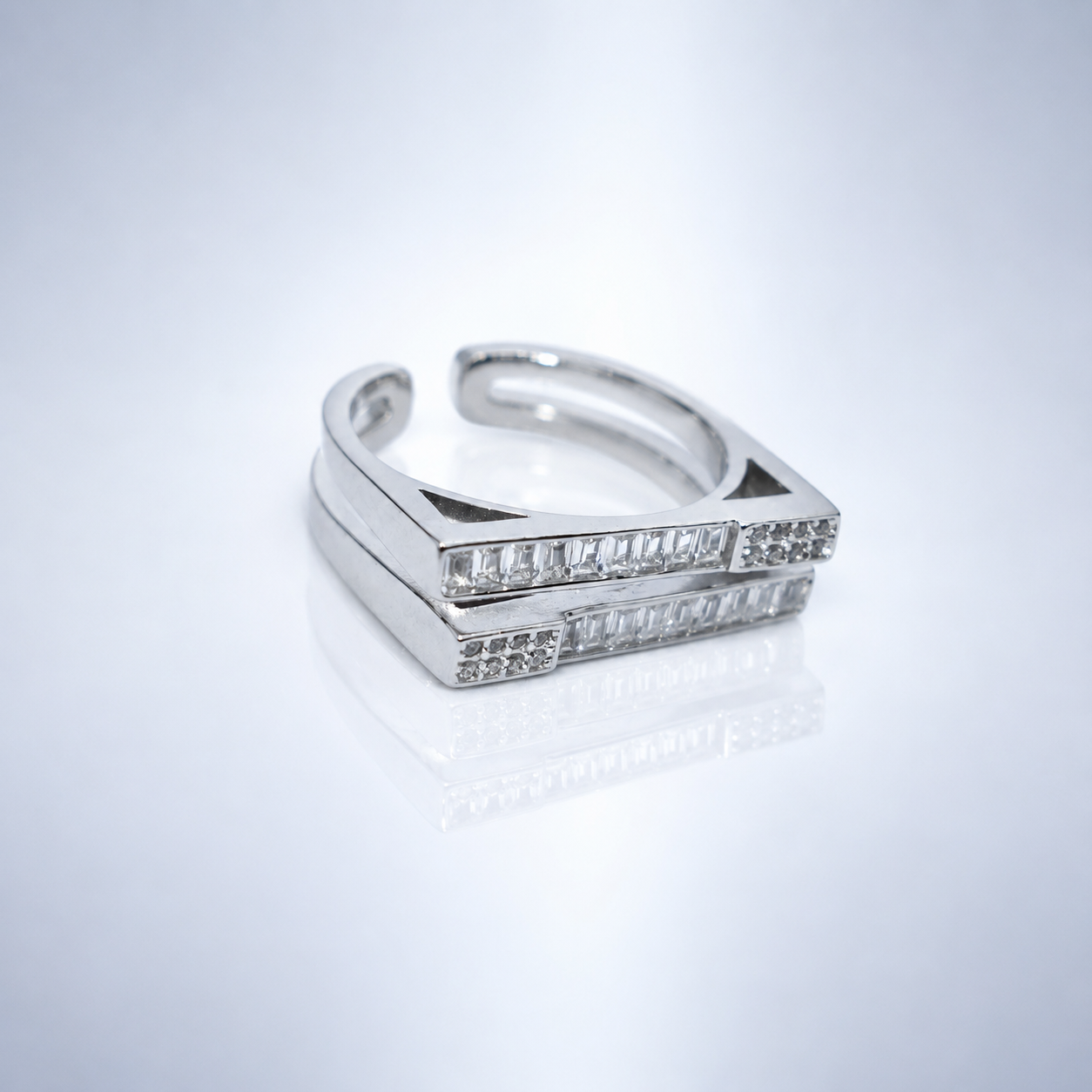 A silver-toned open band ring featuring two parallel rectangular bars set with clear baguette gemstones, photographed on a clean white reflective surface.