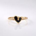A gold ring featuring a heart-shaped bezel-set black enamel center, with a pavé band of small clear stones, photographed on a clean white reflective surface.