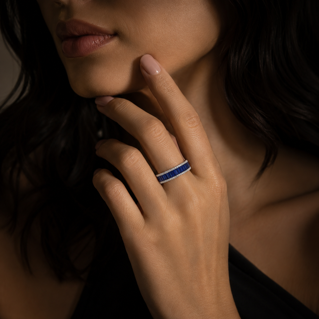 A close-up lifestyle shot of a woman wearing a slim silver band ring set with sparkling blue stones, resting her chin on her hand.