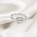 A silver heart-shaped ring with a pavé-set border and a polished silver heart in the center, resting on draped white satin fabric.