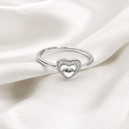 A silver heart-shaped ring with a pavé-set border and a polished silver heart in the center, resting on draped white satin fabric.