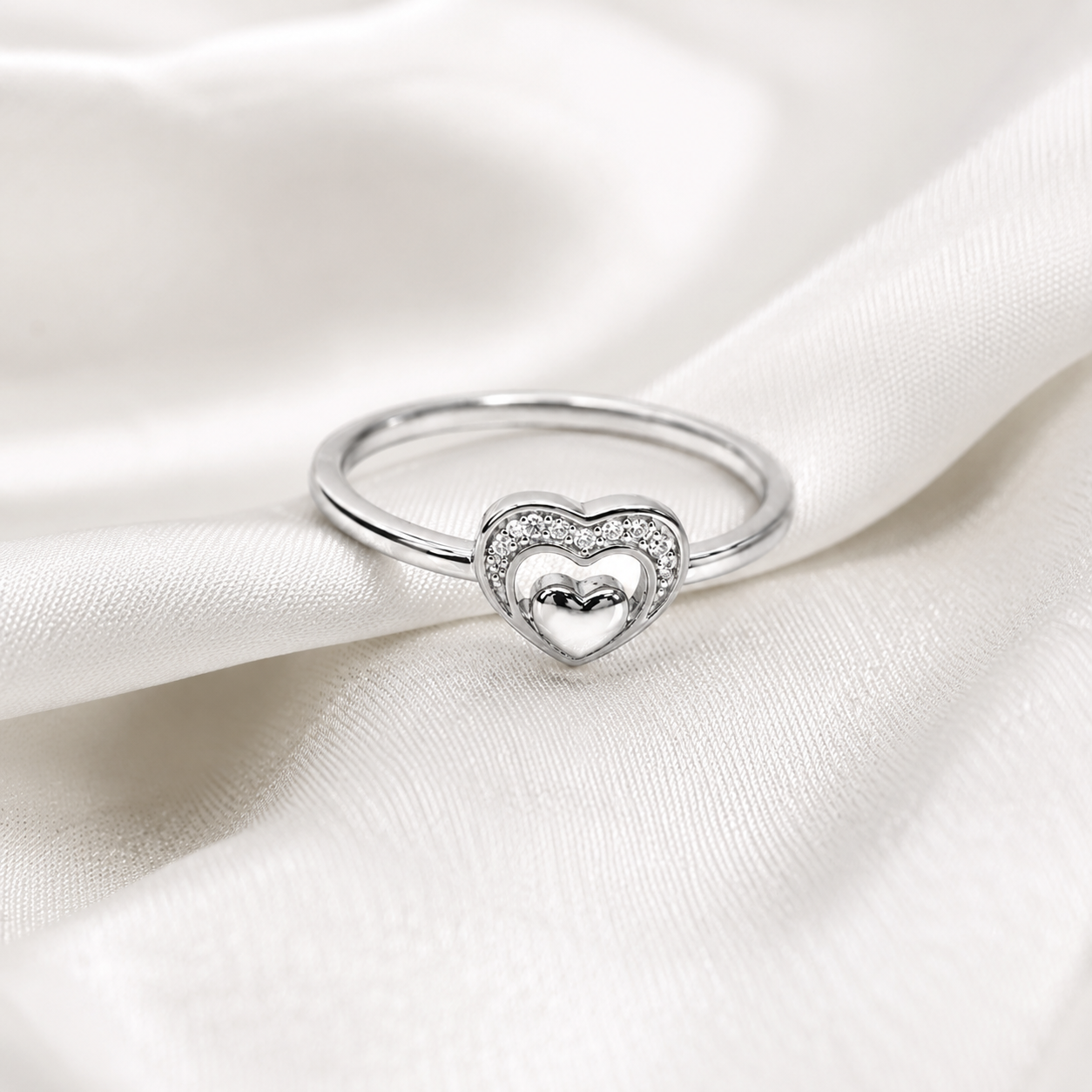 A silver heart-shaped ring with a pavé-set border and a polished silver heart in the center, resting on draped white satin fabric.