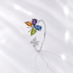 A silver butterfly ring featuring four colorful gemstones in teardrop shapes (orange, purple, green, and blue) and a smaller clear stone butterfly below, set against a white background with a soft reflection.