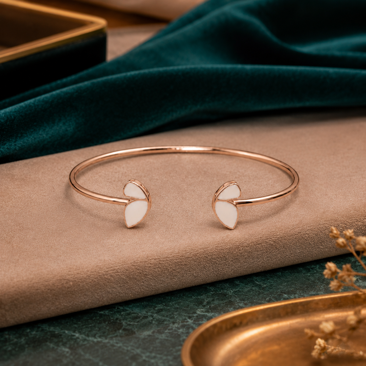 A minimalist rose gold open cuff bangle with two small white enamel leaf-like accents, displayed on a beige textured surface with a dark green velvet cloth in the background.
