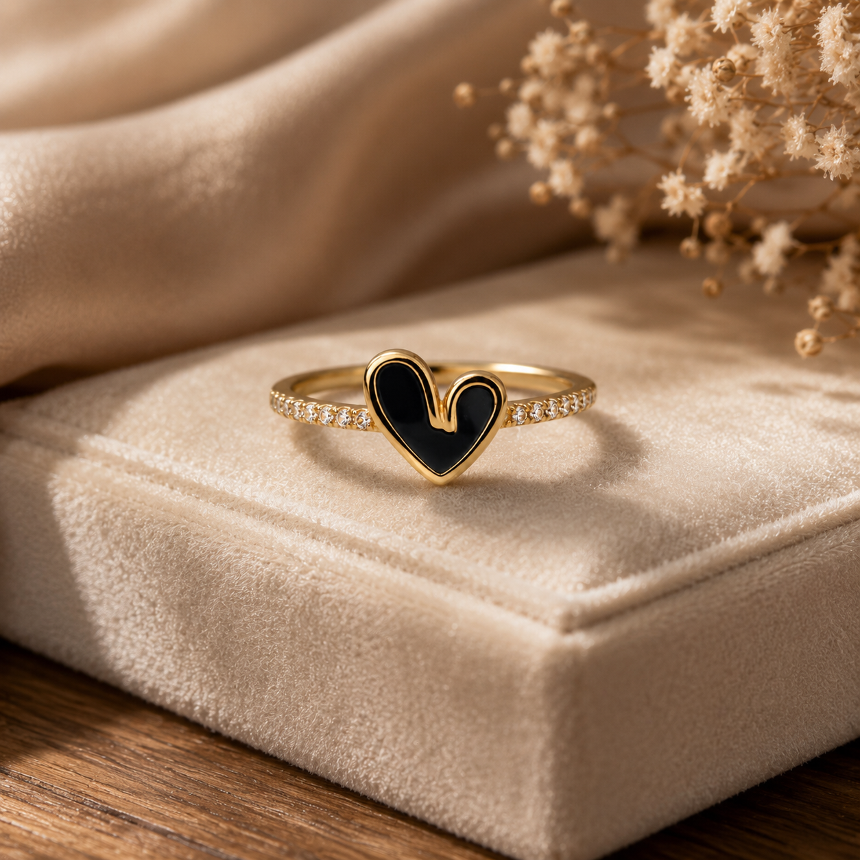 A gold heart-shaped ring with a black enamel center and small clear stones along the band, displayed on a beige velvet jewelry box with dried flowers in the background.