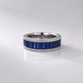A silver band ring featuring a row of deep blue baguette-cut stones bordered by small clear stones, photographed on a clean light gray reflective surface.