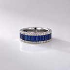 A silver band ring featuring a row of deep blue baguette-cut stones bordered by small clear stones, photographed on a clean light gray reflective surface.