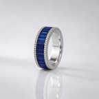A silver band ring featuring a central channel of deep blue baguette-cut gemstones, bordered by a halo of small clear stones, shown on a reflective light gray surface.
