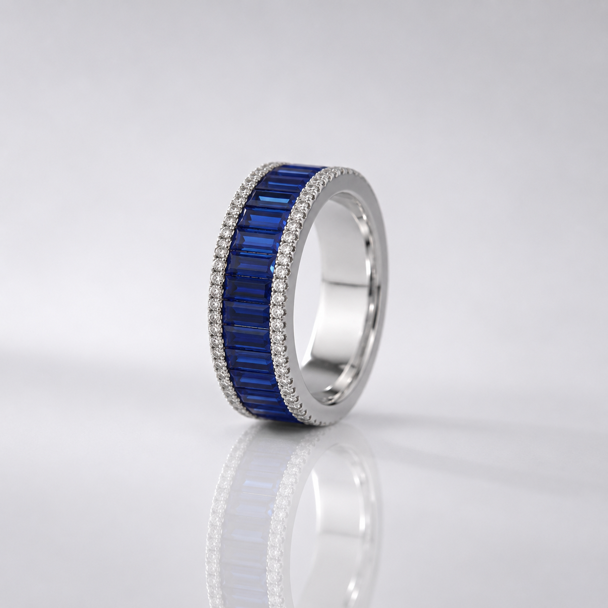 A silver band ring featuring a central channel of deep blue baguette-cut gemstones, bordered by a halo of small clear stones, shown on a reflective light gray surface.