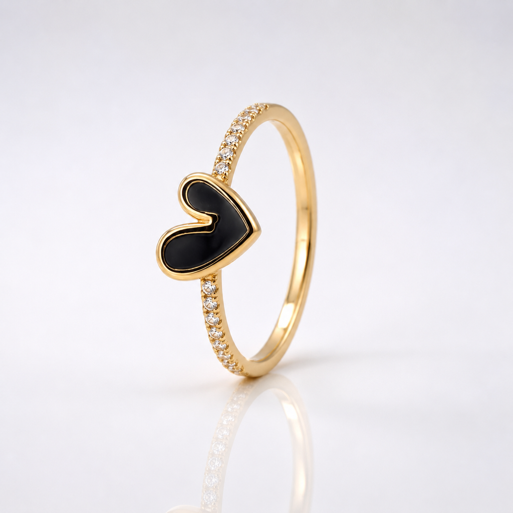 A gold ring featuring a central heart-shaped inlay with black enamel, accented by small clear stones along the band, set against a clean white background with a soft reflection.
