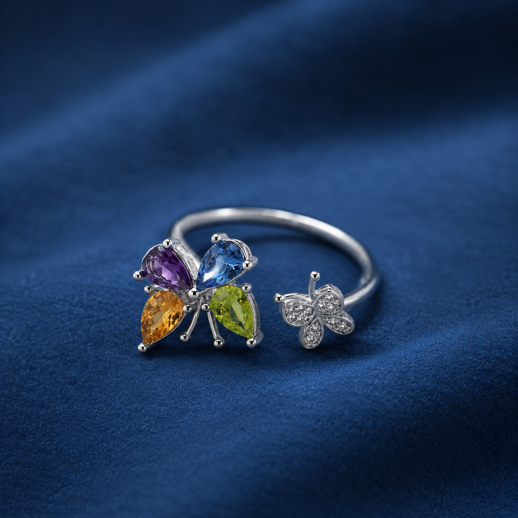A silver butterfly ring set with multicolored gemstones, displayed on a dark blue fabric background. The butterfly head features four marquise-cut gemstones in purple, blue, yellow-green, and yellow, while the wing is adorned with small clear cubic zirconia stones.