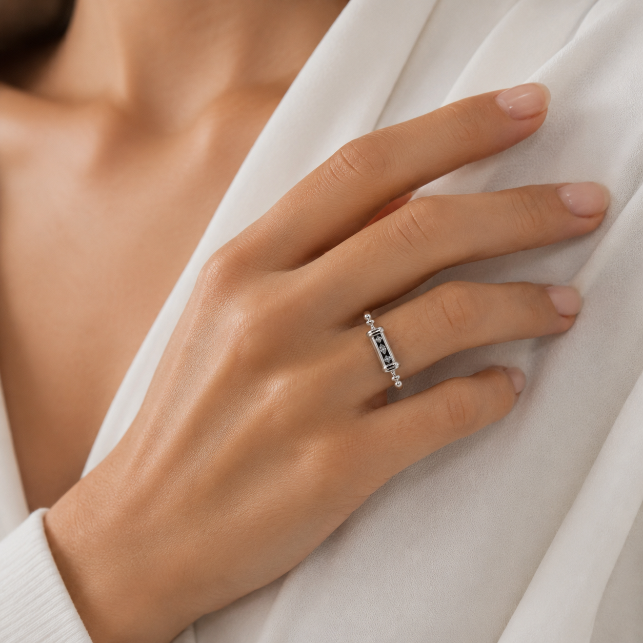 A close-up of a hand wearing a delicate silver ring with a central black-and-white patterned inlay, resting against white draped fabric.