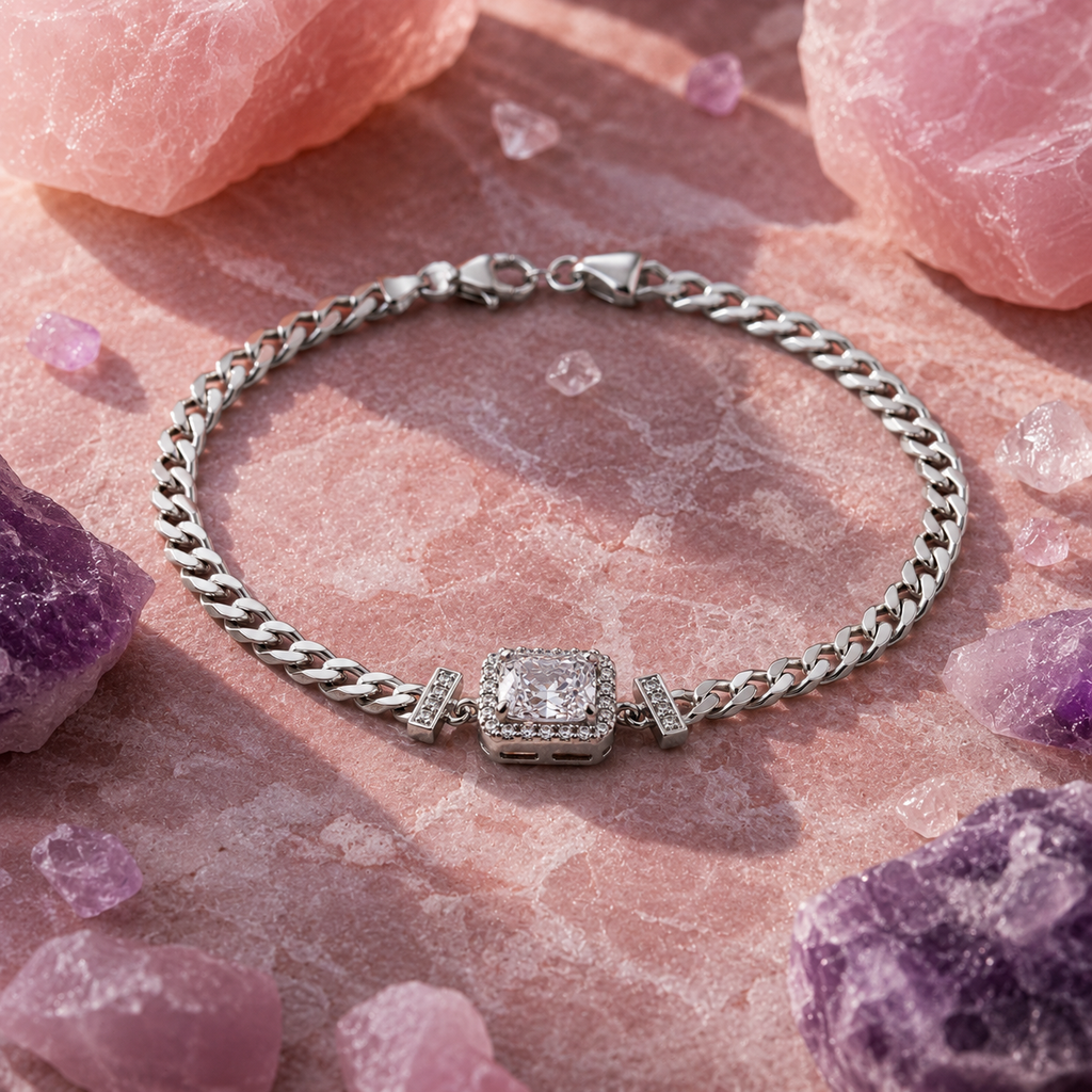 A silver-toned curb chain bracelet with a central square halo-set clear gemstone, displayed on a pink stone surface surrounded by pink and purple crystals.