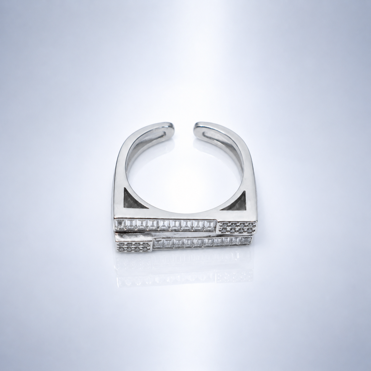 A silver-toned geometric ring with a wide curved band and rows of small clear stones, photographed on a clean light gray reflective surface.