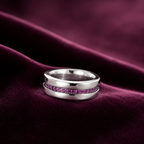 A polished silver ring with a channel-set row of small purple gemstones, displayed on draped dark burgundy velvet fabric.