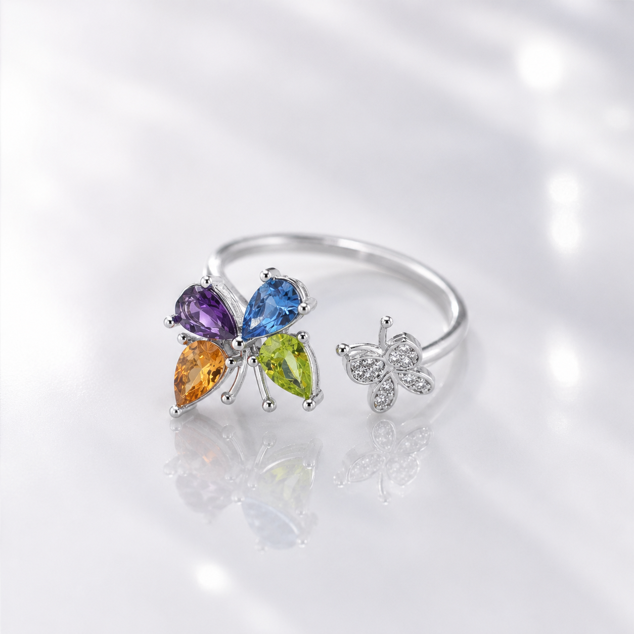 A silver butterfly ring set with four colorful gemstones (purple, blue, green, and orange) and a smaller matching butterfly charm, displayed on a reflective white surface.