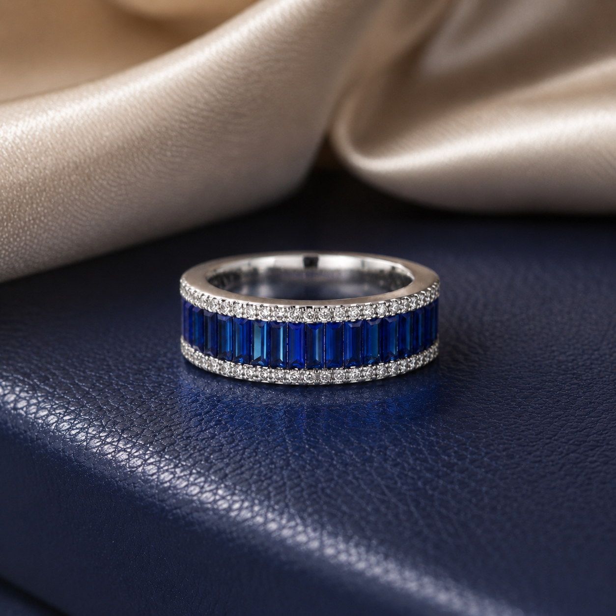 A silver band ring featuring two rows of rectangular deep blue gemstones and small clear stones, displayed on a textured navy surface with draped champagne satin in the background.