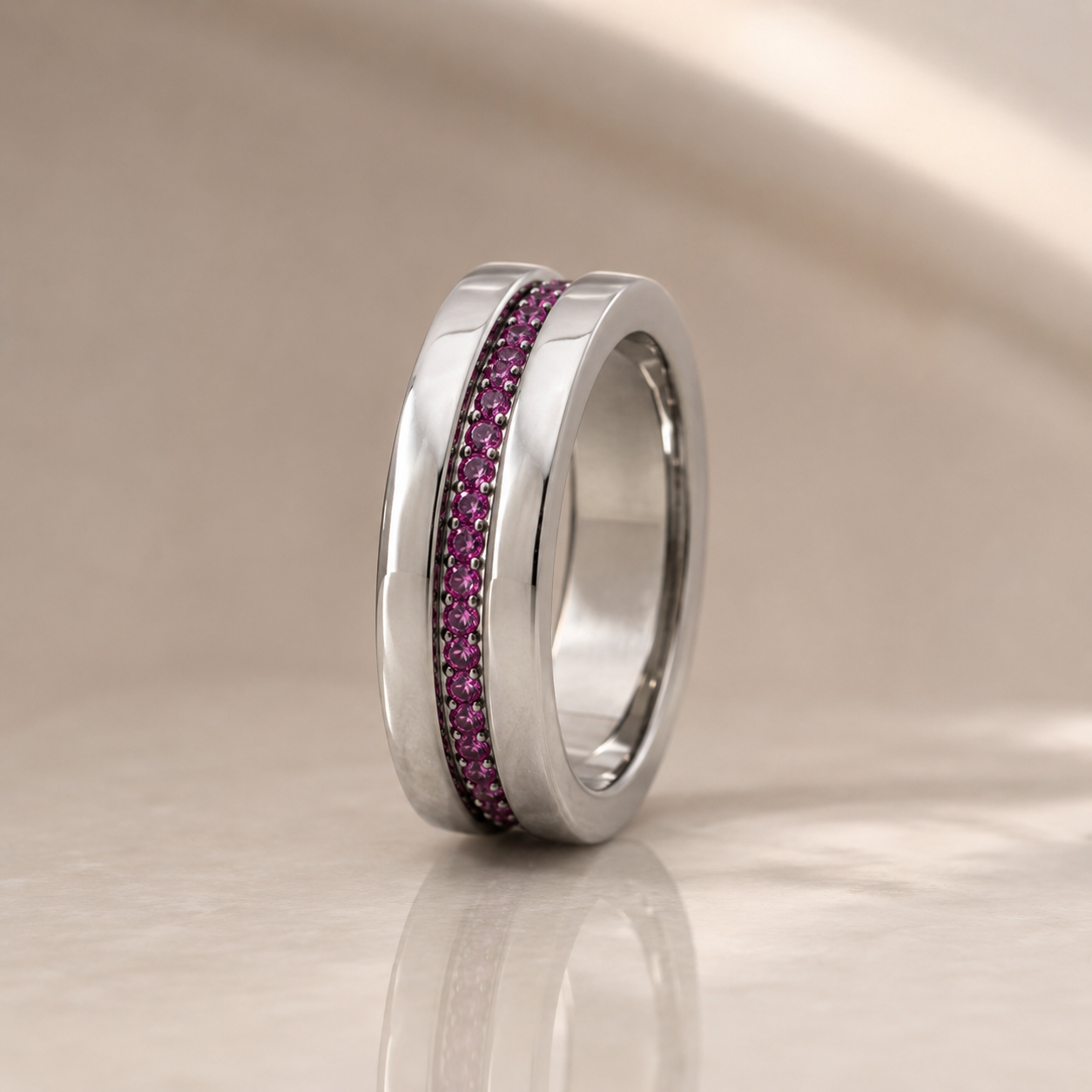 A polished silver-tone ring featuring a central channel set with small pink gemstones, displayed on a reflective beige surface.