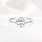 A silver heart-shaped ring with a polished center heart and a halo of small clear stones on the band, photographed on a soft white background with a faint reflection below.