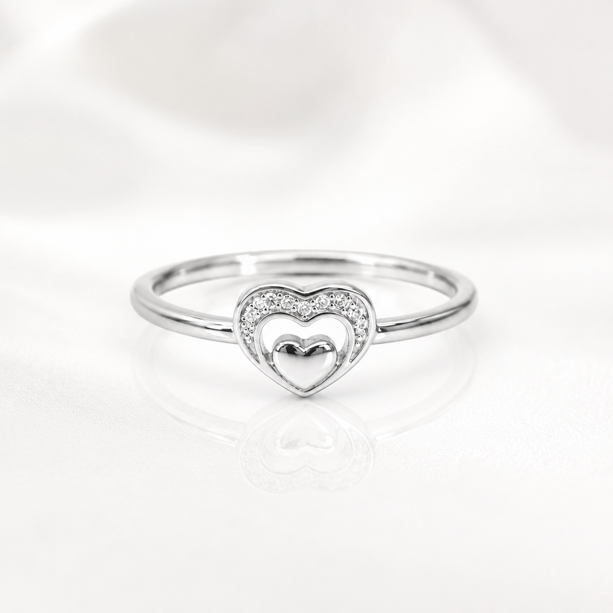A silver heart-shaped ring with a polished center heart and a halo of small clear stones on the band, photographed on a soft white background with a faint reflection below.