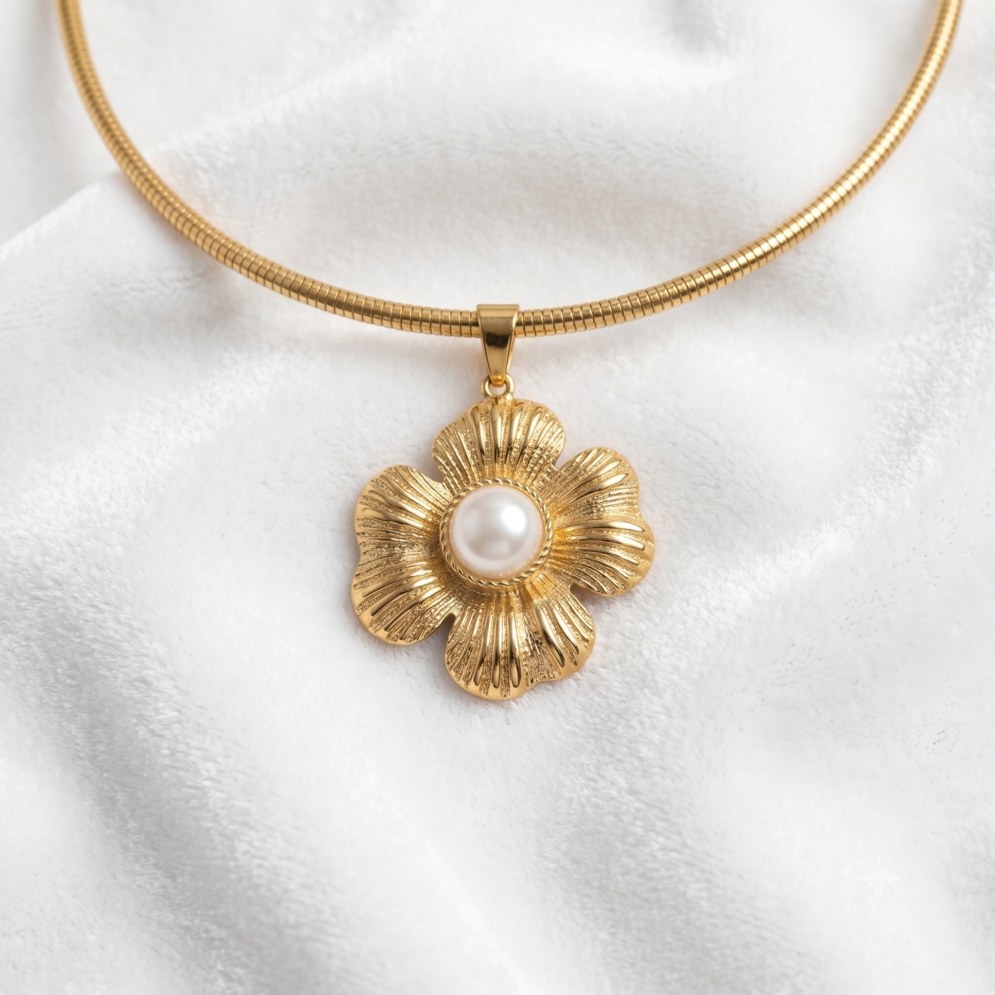 A gold snake-chain necklace with a textured gold flower pendant featuring a white faux-pearl center, displayed on a soft white fabric background.