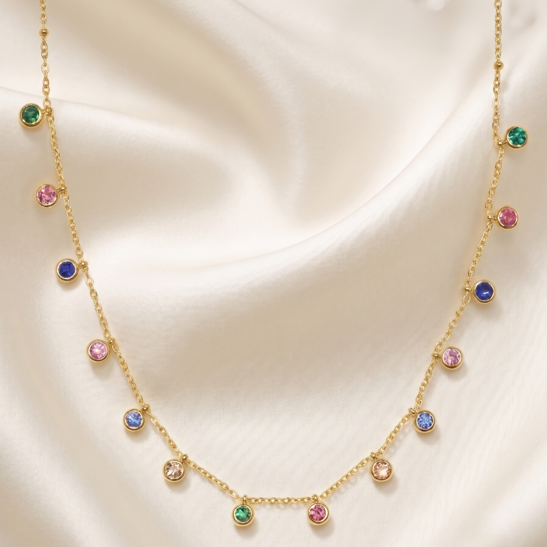 A delicate gold chain necklace draped elegantly on a soft, cream-colored satin fabric. The necklace features multiple small, round bezel-set gemstones in various colors including green, pink, blue, and purple, set in gold.