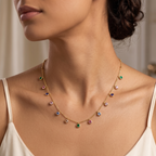 A close-up lifestyle shot of a model wearing a delicate gold necklace adorned with small, colorful gemstone drops in green, pink, blue, and purple.